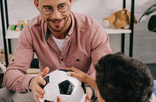 smiling teacher and african american preschooler holding soccer ball together in classroom